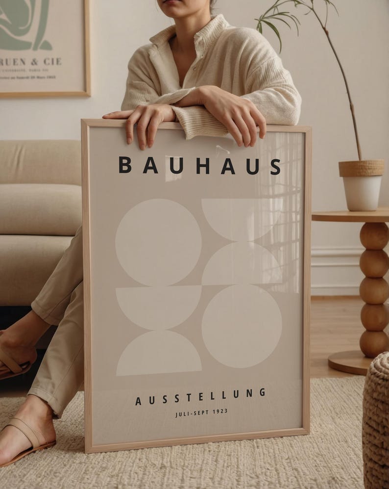 May include: A framed Bauhaus exhibition poster. The poster features a beige background with white geometric shapes and the word "BAUHAUS" in black. Below, the text "AUSSTELLUNG" and "JULI-SEPT 1923" are also in black. The frame is light brown.