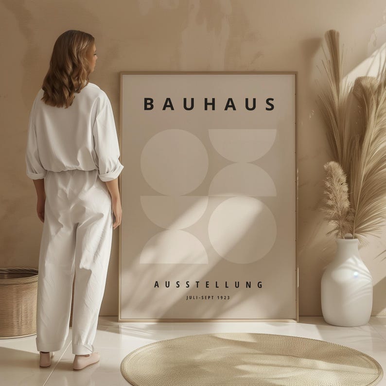 May include: A framed Bauhaus exhibition poster with a beige background and geometric shapes. The poster features the word "BAUHAUS" at the top and "AUSSTELLUNG JULI-SEPT 1923" at the bottom. A person in white clothing stands next to the poster.