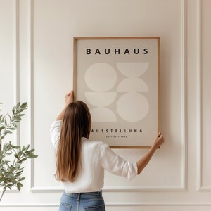 May include: A framed Bauhaus exhibition poster with a beige background and geometric shapes. The poster is held by a person in a white shirt and blue jeans. The poster's text reads "BAUHAUS AUSSTELLUNG 1923-1923".