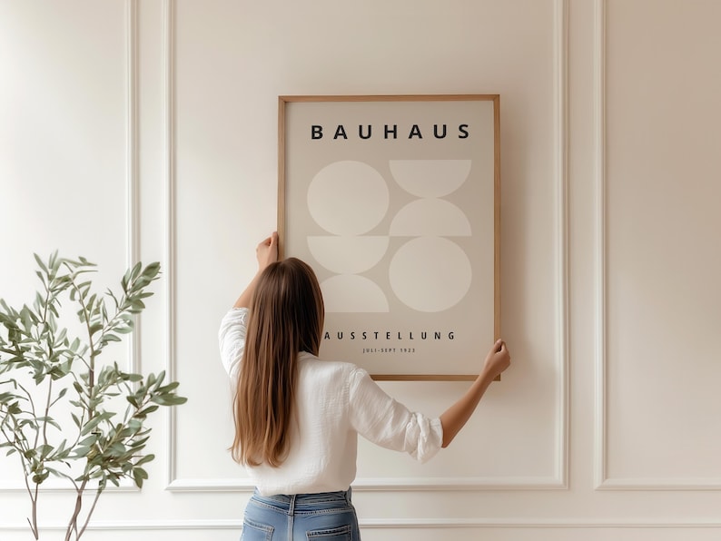 May include: A framed Bauhaus exhibition poster with a beige background and geometric shapes. The poster is held by a person in a white shirt and blue jeans. The poster's text reads "BAUHAUS AUSSTELLUNG 1923-1923".