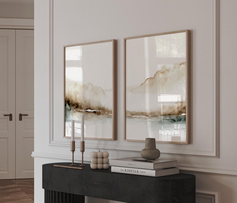 May include: Two framed abstract landscape paintings with brown and blue watercolor washes, displayed on a black console table with candles, a decorative object, and a stack of books. The top book has the text "The Kinfolk Table" on the cover.
