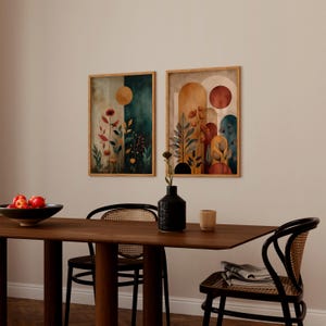 Puede incluir: Dos impresiones art&iacute;sticas bot&aacute;nicas enmarcadas. Una presenta un fondo verde oscuro y beige con un c&iacute;rculo dorado y flores rojas. La otra tiene un fondo beige y naranja con un c&iacute;rculo rojo y elementos florales. Las impresiones se exhiben sobre una mesa de madera.