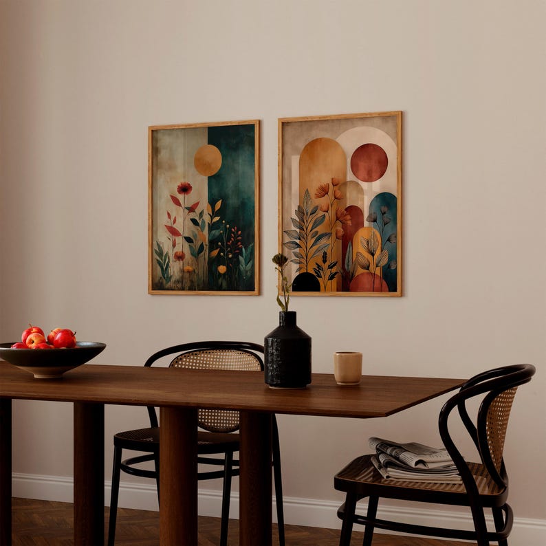 Puede incluir: Dos impresiones art&iacute;sticas bot&aacute;nicas enmarcadas. Una presenta un fondo verde oscuro y beige con un c&iacute;rculo dorado y flores rojas. La otra tiene un fondo beige y naranja con un c&iacute;rculo rojo y elementos florales. Las impresiones se exhiben sobre una mesa de madera.
