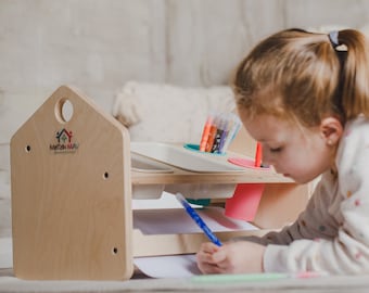 Può includere: Una scrivania artistica in legno per bambini con un pannello laterale a forma di casa. La scrivania ha un vassoio integrato per materiali artistici e un ripiano inferiore per la carta. Il bambino è sdraiato sul pavimento, che disegna con una penna blu. La scrivania ha il testo "Matak-Matak".