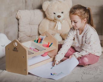 May include: A child sits on a rug with a world map design, drawing on a piece of paper. The child is using a wooden art easel with three compartments for supplies. The easel has a white surface for drawing and a label that says "Make & Learn".