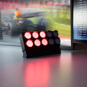 Könnte beinhalten: Eine schwarze, rechteckige Rennflaggen-Lichtanzeige mit zwei Reihen von vier kreisförmigen Lichtern. Die obere Reihe zeigt drei leuchtende rote Lichter und ein unbeleuchtetes Licht, während die untere Reihe drei leuchtende rote Lichter und zwei unbeleuchtete Lichter zeigt.