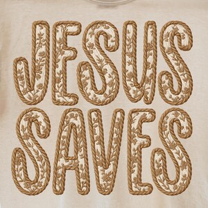 May include: Cream-colored t-shirt with the words "JESUS SAVES" in a decorative, rope-like font. The letters are filled with a floral pattern. The shirt is hanging on a wooden hanger.