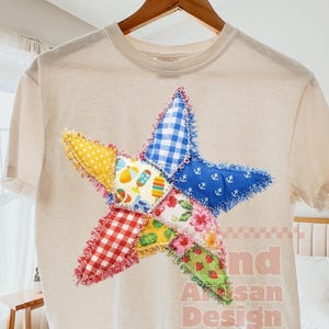 Puede incluir: Camiseta beige con una gran estrella de patchwork colorida. La estrella está hecha de varios patrones de tela, incluyendo cuadros, estampados florales y náuticos. El texto "nd Artisan Design" está impreso en la camiseta.