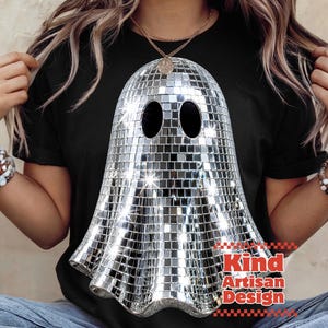 May include: A black t-shirt featuring a large graphic of a ghost made of reflective silver disco ball mirror tiles. The ghost has two oval eye cutouts. The text Kind Artisan Design is printed in red letters near the bottom right of the graphic.