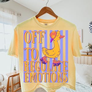 Puede incluir: Camiseta amarilla clara con la frase "OFF TO REGULATE EMOTIONS" en naranja y morado. El diseño presenta un pato amarillo con sombrero y patines. La camiseta está colgada de una percha de madera.