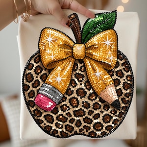 May include: A cream-coloured t-shirt featuring a large, circular sequined applique of an apple with a leopard print pattern. The apple is adorned with a prominent gold sequined bow, a green sequined leaf, and a pencil design with a pink eraser.