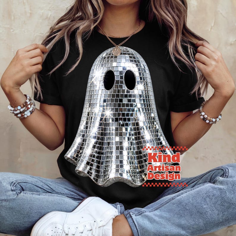 May include: A black t-shirt featuring a large graphic of a ghost made of reflective silver disco ball mirror tiles. The ghost has two oval eye cutouts. The text Kind Artisan Design is printed in red letters near the bottom right of the graphic.