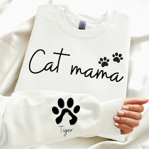 May include: White sweatshirt with the text "Cat mama" and two paw prints. The left sleeve has a paw print with a cat silhouette inside and the text "Tiger" underneath.