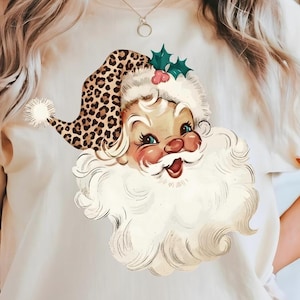 May include: A cream-coloured t-shirt featuring a graphic illustration of a Santa Claus face with a large white beard and a leopard print hat. The hat is accented with a white pom-pom and a small sprig of green holly leaves with red berries.