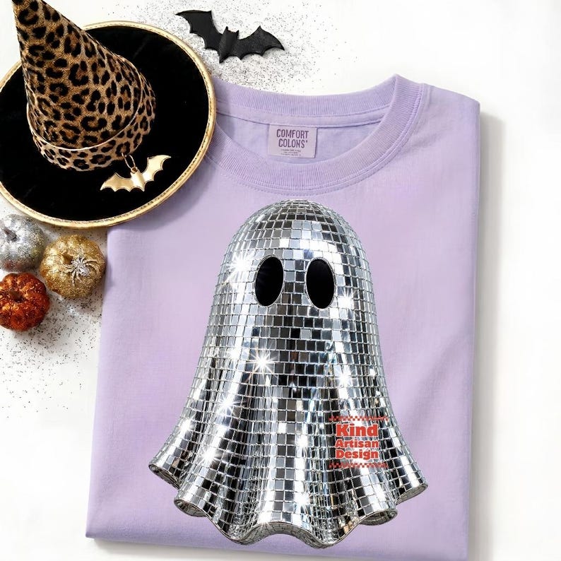 May include: A light purple t-shirt featuring a graphic of a ghost made of silver mirror tiles. The shirt has a Comfort Colors label. Nearby is a leopard print witch hat with a black brim, a black bat cutout, and three small decorative pumpkins.
