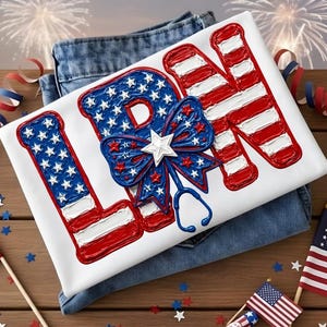 May include: A white t-shirt featuring the letters LPN in a raised, textured American flag pattern. The design includes a blue bow with white stars and a stethoscope outline integrated into the lettering, all rendered in red, white, and blue colours.