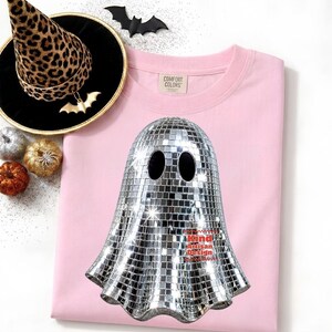May include: A light pink t-shirt featuring a graphic of a ghost covered in silver mirror tiles, resembling a disco ball. The shirt includes a Comfort Colors label. Nearby is a leopard print witch hat, a black bat cutout, and small pumpkins.