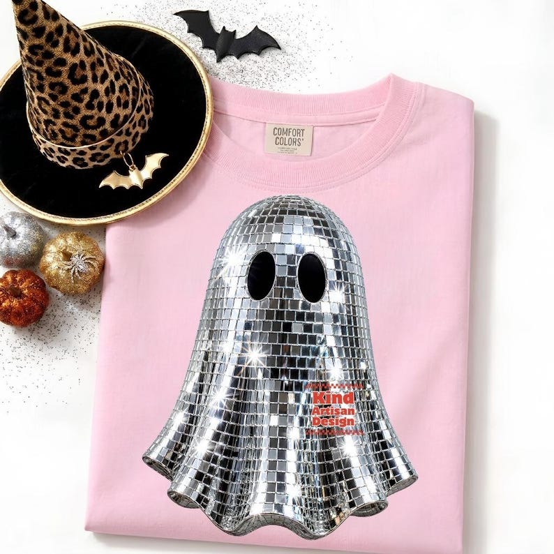May include: A light pink t-shirt featuring a graphic of a ghost covered in silver mirror tiles, resembling a disco ball. The shirt includes a Comfort Colors label. Nearby is a leopard print witch hat, a black bat cutout, and small pumpkins.