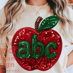 Può includere: Una t-shirt color crema con una grande grafica centrale a forma di mela realizzata con paillettes rosse. La mela ha una foglia e un gambo di paillettes verdi, con le lettere minuscole 'abc' in paillettes verdi al centro del disegno.