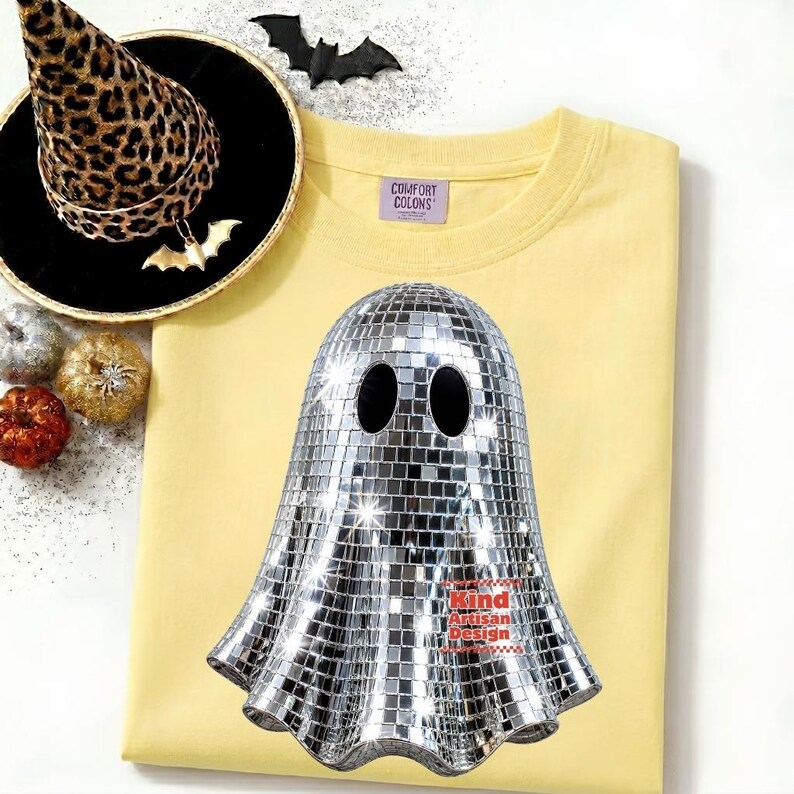 May include: A pale yellow t-shirt featuring a graphic of a ghost made of silver mirrored disco ball tiles. The ghost has two black oval eyes. The shirt has a Comfort Colors label. Nearby are a leopard print witch hat and small pumpkins.