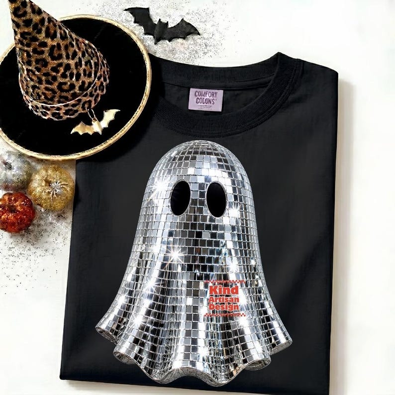 May include: A black t-shirt featuring a silver disco ball ghost graphic with two oval eye cutouts. The shirt includes a Comfort Colors label. Accompanying items include a leopard print witch hat, a black bat cutout, and small decorative pumpkins.