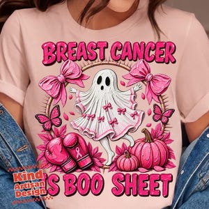 Puede incluir: Una camiseta rosa claro con un gráfico de un fantasma blanco adornado con pequeños lazos rosas, rodeado de guantes de boxeo rosas con un símbolo de cinta, calabazas rosas, mariposas y grandes lazos rosas. El texto dice BREAST CANCER IS BOO SHEET.