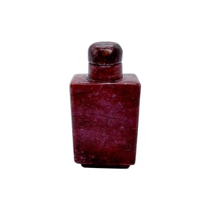 May include: A small, rectangular, dark red bottle with a rounded top and a matching stopper. The bottle has a textured surface and appears to be made of stone or a similar material. The stopper is a darker shade of red.