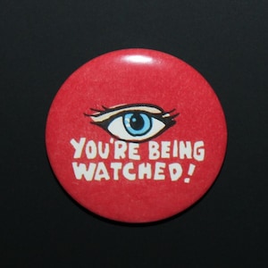 May include: A circular red pin-back button featuring a graphic illustration of a single blue eye with black eyelashes centered above the white text that reads YOU'RE BEING WATCHED! in a bold, hand-drawn style font on the red background.