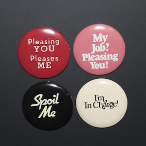 May include: Four circular pin-back buttons with text. The top left is dark red with white text reading Pleasing YOU Pleases ME. The top right is pink with white text reading My Job? Pleasing You! The bottom left is black with white text reading Spoil Me. The bottom right is off-white with black text reading I'm In Charge!