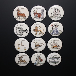 May include: A set of twelve circular white ceramic tiles, each featuring a hand-drawn illustration of a zodiac sign in black ink with subtle color accents. The signs include Aries, Taurus, Gemini, Cancer, Leo, Virgo, Libra, Scorpio, Sagittarius, Capricorn, Aquarius, and Pisces, each labeled with its name in a gothic-style font.