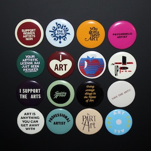 May include: A collection of sixteen circular pin-back buttons arranged in a four-by-four grid. Each button features unique text and graphic designs in various colors, including phrases like Support Women Artists Now, I Heart Art, and Professional Artist.