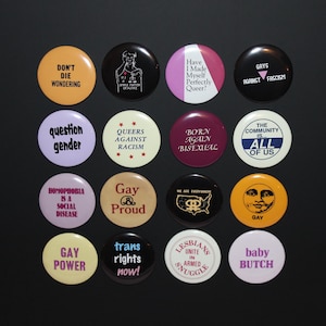 May include: A collection of sixteen circular pin-back buttons arranged in a four-by-four grid. Each button features unique text and graphic designs in various colors, including yellow, black, white, purple, maroon, and cream, with political and social slogans.