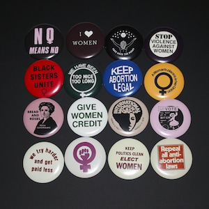 May include: A collection of sixteen circular pinback buttons arranged in a four-by-four grid. Each button features unique text and graphic designs related to social and political activism, including slogans like No Means No, Black Sisters Unite, and Give Women Credit.