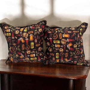 May include: Two black throw pillows with a repeating pattern of beer mugs, pretzels, hops, and other beer-related imagery. The pillows are on a dark wooden surface.