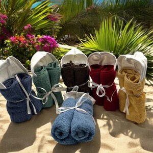 May include: Six hooded towels in different colors, including blue, green, brown, red, yellow, and a light blue color. The towels are folded and tied with white ribbon. The towels are on a sandy beach.