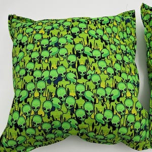 May include: Green pillow with a repeating pattern of green aliens. The aliens are all facing forward and have large eyes and small mouths.