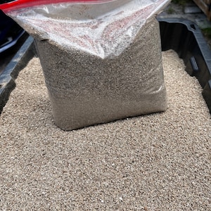 May include: A clear plastic bag filled with small, light brown and white gravel. The bag is sitting on a larger pile of the same gravel.