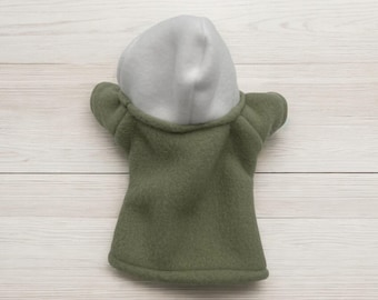 May include: A green fleece hooded puppet with a grey hood. The puppet is lying on a white wooden surface.