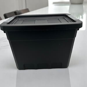 May include: A black plastic storage bin with a lid and two handles. The bin has a textured surface and is sitting on a white surface.