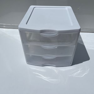 May include: A white plastic storage unit with three clear drawers and a top lid. The unit is designed for organizing small items. The drawers have curved handles for easy opening and closing. The unit is sitting on a white surface.
