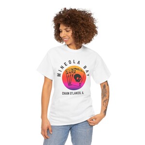 以下が含まれることがあります： 白いTシャツ。円形のデザインで、「MINEOLA BAY」と「CHAIN O'LAKES, IL」の文字が入っています。オレンジ、ピンク、紫のグラデーションの背景に、トランプとスカルが描かれています。