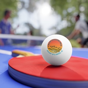 May include: A white ping pong ball with the text "I Hate Pulling Out" in a circular design with a boat graphic. The ball is resting on a red ping pong paddle on a blue table.