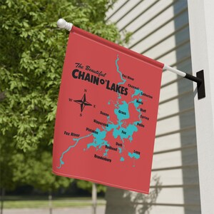 Puede incluir: Una bandera roja con un mapa de Chain O' Lakes, con el texto "The Beautiful Chain O' Lakes". El mapa muestra los lagos en azul, con nombres como Fox River y Pistakee. También se presenta una rosa de los vientos.