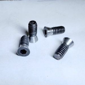 May include: Four black metal screws with hexagonal heads and a countersunk design. The screws are arranged on a white surface.