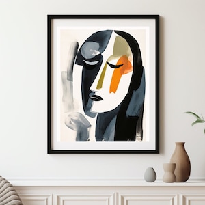 May include: Abstract art print depicting a stylized face with closed eyes. The artwork uses a palette of black, white, grey, orange, and olive green. The print is framed in a black frame and displayed on a white wall.
