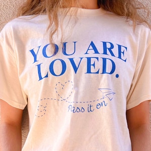 May include: A white t-shirt with blue text that reads "You Are Loved." A blue paper airplane and heart are drawn below the text with the words "Pass it on" underneath.