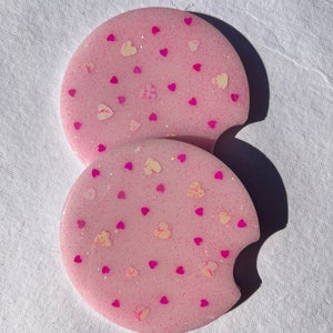 May include: Two pink round coasters with a pink glitter finish and a scattered pattern of small pink and white hearts.