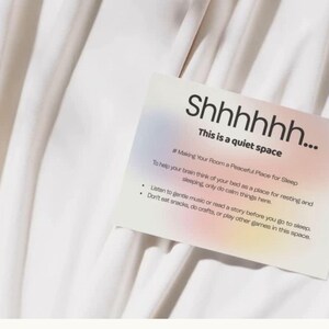 May include: A white card with the text "Shhhhhh... This is a quiet space" on a white fabric background. The card includes instructions for creating a peaceful sleep environment, such as listening to music and avoiding snacks.