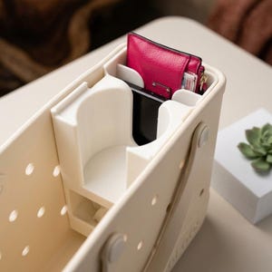 May include: A cream-colored plastic storage basket with a perforated side design and a built-in white interior organizer featuring a cup holder and slots. A bright pink textured wallet and a black rectangular object are tucked into the organizer.