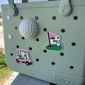 May include: A light green tote bag adorned with golf-themed accessories. A white golf ball and three golf-themed stickers are attached to the bag. The stickers include a golf cart, a golf ball on a green, and a pink flag with the words "Hole in One".